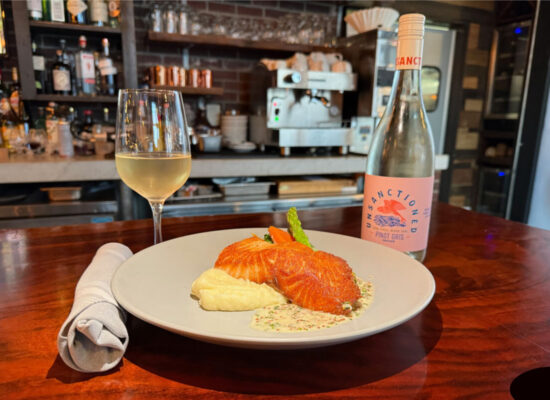 plate of pan-roasted salmon with whipped potatoes and market grilled vegetables on a wooden bar. A glass of white wine to the left and wine bottle to the right of the plate. Roll of silverware in a linen napkin to the left side.